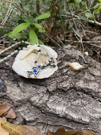 A rosary with a white cross and beads rests on a white stone-like surface placed on a textured tree bark. Green leaves and branches surround the scene, giving a natural and serene ambiance. Fallen brown leaves are scattered around, enhancing the rustic feel.