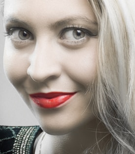 A close-up of a smiling person with bright red lipstick and light-colored eyes, showcasing smooth skin and platinum blonde hair. The focus is on the facial features, highlighting dramatic eye makeup and a vivid smile. The background is neutral, drawing attention to the person's expression.