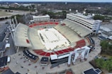 An aerial view of a large outdoor stadium with red and beige seating areas. A central area appears to be set up for an event, possibly with a temporary platform or stage. Surrounding the stadium are multiple buildings and parking areas. Light poles and trees are visible around the venue, with a broader landscape of greenery and roads stretching into the background.