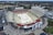 An aerial view of a large outdoor stadium with red and beige seating areas. A central area appears to be set up for an event, possibly with a temporary platform or stage. Surrounding the stadium are multiple buildings and parking areas. Light poles and trees are visible around the venue, with a broader landscape of greenery and roads stretching into the background.