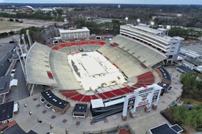 An aerial view of a large outdoor stadium with red and beige seating areas. A central area appears to be set up for an event, possibly with a temporary platform or stage. Surrounding the stadium are multiple buildings and parking areas. Light poles and trees are visible around the venue, with a broader landscape of greenery and roads stretching into the background.