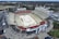 An aerial view of a large outdoor stadium with red and beige seating areas. A central area appears to be set up for an event, possibly with a temporary platform or stage. Surrounding the stadium are multiple buildings and parking areas. Light poles and trees are visible around the venue, with a broader landscape of greenery and roads stretching into the background.