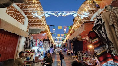 A vibrant market street at dusk with a sky overhead transitioning to twilight. Stalls line both sides of the path, displaying colorful textiles, clothing, and other goods. Bright string lights adorn the top, creating an inviting, festive atmosphere. People stroll along the street, engaging with vendors.