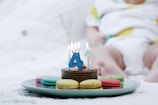 A cake with a blue number 4 candle and several colorful macarons on a green plate. The background features a blurred child sitting down, wearing a white outfit with colorful patterns.