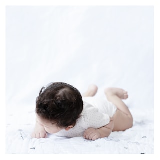 A baby lying on its stomach on a light-colored blanket, with dark curly hair and wearing a white onesie with small patterns.