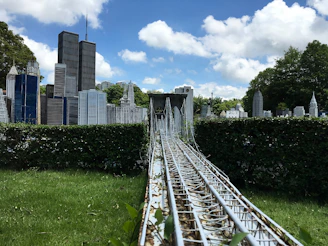 A detailed miniature model of a cityscape features iconic skyscrapers surrounded by greenery. A metal structure resembling a suspension bridge runs through the foreground, leading towards the buildings.