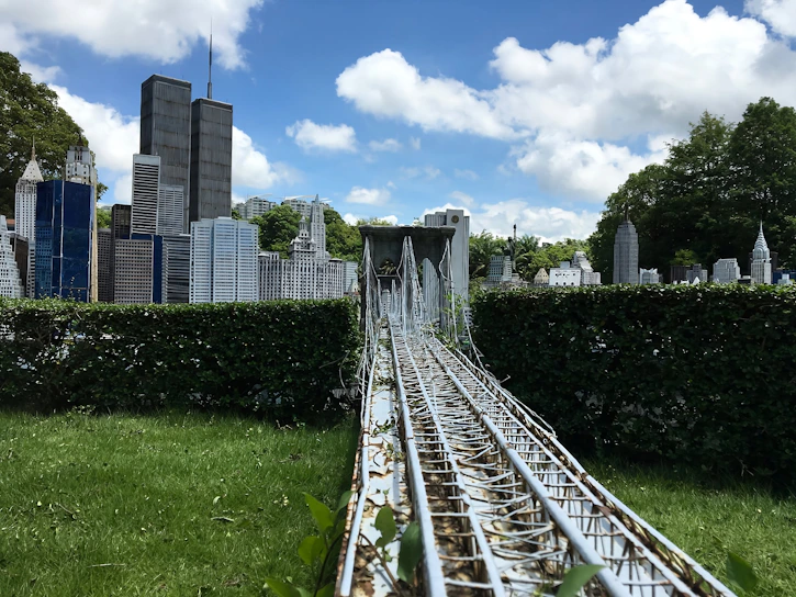 A detailed miniature model of a cityscape features iconic skyscrapers surrounded by greenery. A metal structure resembling a suspension bridge runs through the foreground, leading towards the buildings.