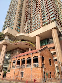 A large modern high-rise building towers above a smaller, historic brick building featuring arched windows and a tiled roof. The contrast between the two architectural styles is striking, with the bright, glass-clad tower juxtaposed against the warm, earthy tones of the brick structure.