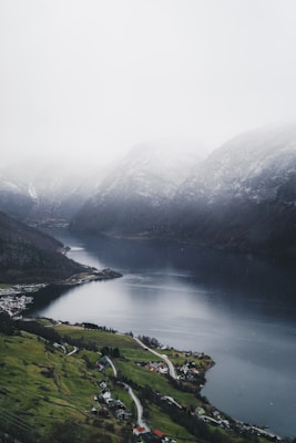 A serene landscape featuring a winding river or fjord surrounded by steep, mist-covered mountains. In the foreground, a small village with scattered houses and green fields is visible, nestled along a narrow road. The sky appears overcast, contributing to a tranquil and contemplative atmosphere.