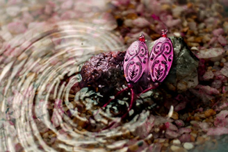 A pair of earrings crafted with rough-cut garnets, displayed on driftwood with ocean waves blurred in the background.