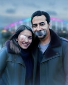 A man and a woman dressed warmly are standing close together, smiling. The woman has a bandage over her right eye with a hint of blood near it, while the man has duct tape across his mouth. In the background, a brightly lit, colorful bridge spans across the image, with blurred lights creating a soft bokeh effect.