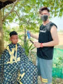 A barber wearing a black sleeveless shirt and mask is holding a spray can and a brush, standing beside a young boy seated for a haircut. The boy is wearing a cape with a designer pattern. They are outdoors with green trees and foliage in the background.