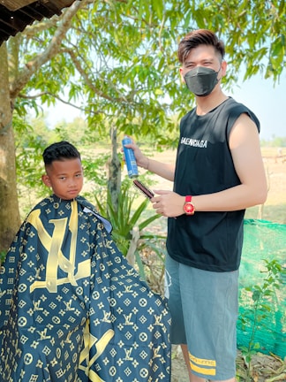 A barber wearing a black sleeveless shirt and mask is holding a spray can and a brush, standing beside a young boy seated for a haircut. The boy is wearing a cape with a designer pattern. They are outdoors with green trees and foliage in the background.