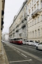 A European city street with a red tram traveling along tracks. The street is lined with ornate, historic buildings with intricate architectural details and balconies. Several cars are parked alongside the street, and the scene has a calm, urban atmosphere.