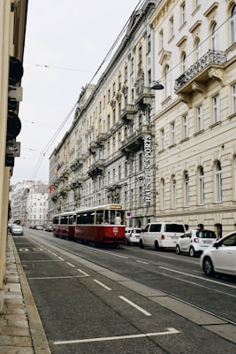 A European city street with a red tram traveling along tracks. The street is lined with ornate, historic buildings with intricate architectural details and balconies. Several cars are parked alongside the street, and the scene has a calm, urban atmosphere.