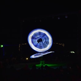 A performer in vibrant, illuminated attire is spinning large, circular, blue and white lights in a dark setting. The light trails create a mesmerizing pattern, captivating the silhouettes of an audience watching the spectacle.