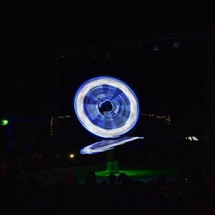 A performer in vibrant, illuminated attire is spinning large, circular, blue and white lights in a dark setting. The light trails create a mesmerizing pattern, captivating the silhouettes of an audience watching the spectacle.