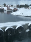 Snow-covered houses with red exteriors sit by a calm body of water. The scene is framed by snow-laden hills and trees, with large black tires lining a concrete barrier at the waterfront.