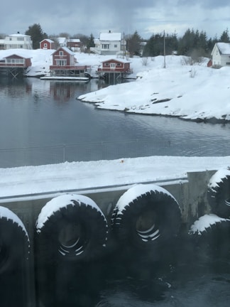 Snow-covered houses with red exteriors sit by a calm body of water. The scene is framed by snow-laden hills and trees, with large black tires lining a concrete barrier at the waterfront.