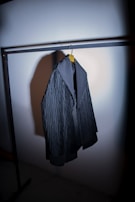 A dark-colored, textured jacket hanging from a clothing rack on a wooden hanger. The jacket has a broad collar and appears to be made of heavy fabric. The lighting is focused, casting a shadow on the wall behind the jacket.