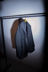 A dark-colored, textured jacket hanging from a clothing rack on a wooden hanger. The jacket has a broad collar and appears to be made of heavy fabric. The lighting is focused, casting a shadow on the wall behind the jacket.