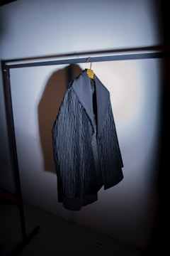 A dark-colored, textured jacket hanging from a clothing rack on a wooden hanger. The jacket has a broad collar and appears to be made of heavy fabric. The lighting is focused, casting a shadow on the wall behind the jacket.