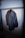 A dark-colored, textured jacket hanging from a clothing rack on a wooden hanger. The jacket has a broad collar and appears to be made of heavy fabric. The lighting is focused, casting a shadow on the wall behind the jacket.