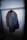 A dark-colored, textured jacket hanging from a clothing rack on a wooden hanger. The jacket has a broad collar and appears to be made of heavy fabric. The lighting is focused, casting a shadow on the wall behind the jacket.