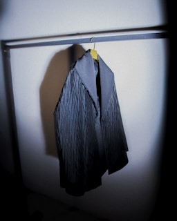 A dark, textured coat hangs on a metal rack. The coat features a subtle pattern and a contrasting collar, positioned in a dimly lit space that casts a shadow on the nearby wall.