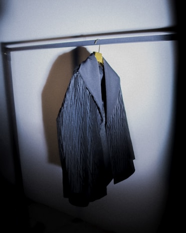 A dark, textured coat hangs on a metal rack. The coat features a subtle pattern and a contrasting collar, positioned in a dimly lit space that casts a shadow on the nearby wall.