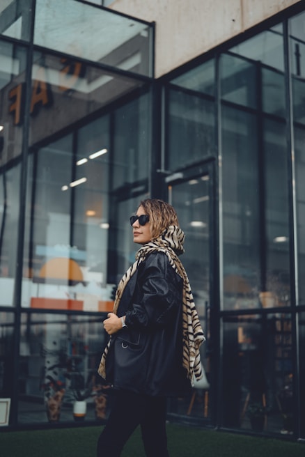 A person wearing sunglasses and a stylish outfit stands in front of a modern glass building. They are wearing a long coat and a striped scarf, exuding a confident, fashionable vibe.