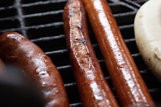 A close-up view of grilled sausages on a grate. The sausages are glistening with oil and are cooked to a golden brown. There are three sausages visible, two darker and one lighter in color.