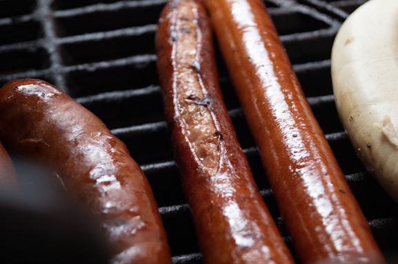 A close-up view of grilled sausages on a grate. The sausages are glistening with oil and are cooked to a golden brown. There are three sausages visible, two darker and one lighter in color.