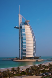 A striking modern architectural structure resembling a sail, located on an artificial island surrounded by turquoise waters and soft, sandy beaches. Lush greenery and palm trees are visible in the foreground.