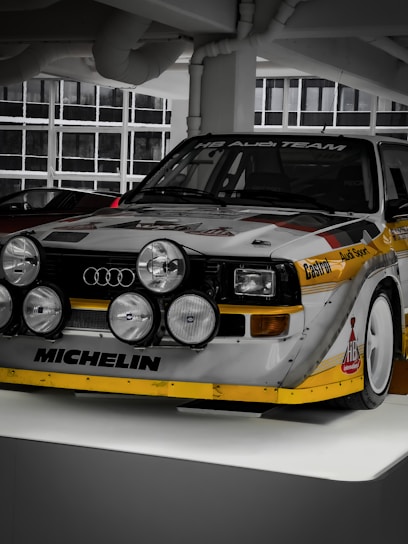 A vintage racing car displayed indoors, prominently featuring the Audi logo and several rally headlights mounted on the front. The vehicle is adorned with sponsor decals such as Michelin and Castrol on a white and yellow color scheme, with large wheel arches and a distinctive retro design.
