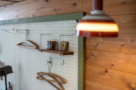 A wooden wall with a pegboard displaying a few items, including a ceramic mug and a small wooden plaque on a shelf. Additional hooks and wooden hangers are mounted on the board, with a hanging lamp providing warm lighting.