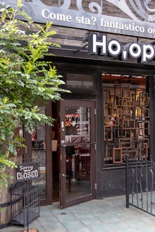 A restaurant entrance features an open glass door with signage displaying opening and closing times. Above the door, a sign reads, 'Come sta? fantastico os' and features decorative scrollwork. A small wooden sign placed outside indicates that the place is closed. The area is adorned with green foliage and metal railings, providing an inviting look.