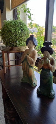 Two decorative ceramic figurines dressed in traditional attire are placed on a dark wooden table. Behind them is a potted round green plant on a small stand, set against a window with a view of a cityscape and greenery outside. The setting suggests an indoor arrangement with natural light coming in through the window.