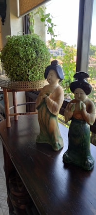 Two decorative ceramic figurines dressed in traditional attire are placed on a dark wooden table. Behind them is a potted round green plant on a small stand, set against a window with a view of a cityscape and greenery outside. The setting suggests an indoor arrangement with natural light coming in through the window.