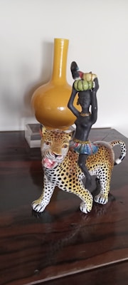 A decorative figurine depicts a dark-skinned figure riding a leopard. The figure carries a basket of fruits on their head and wears a colorful skirt. The leopard is detailed with a spotted pattern and open mouth. In the background is a large, mustard-colored vase on a wooden surface.