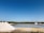 Several large white mounds of salt are located near a calm body of water, with clear blue skies overhead. The landscape is flat, and there is a line of green trees in the distance. The ground appears dry and rocky, with patches of standing water around the salt mounds.