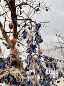 A tree with numerous branches adorned with blue and white amulets, commonly known as evil eye beads. These beads are mostly circular with a central dark spot surrounded by lighter blue and white layers. The tree is bare of leaves, and the sky in the background is overcast.