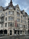 A historic-looking multi-story stone building with numerous windows and a pointed roof. The structure features several architectural details, such as arched windows and turrets. A vertical red sign with the word 'HOTEL' is prominently mounted on the facade. The street in front is wet, suggesting recent rain.