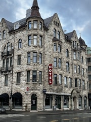 A historic-looking multi-story stone building with numerous windows and a pointed roof. The structure features several architectural details, such as arched windows and turrets. A vertical red sign with the word 'HOTEL' is prominently mounted on the facade. The street in front is wet, suggesting recent rain.