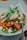 A colorful salad is presented on a plate, featuring fresh ingredients such as sliced tomatoes, leafy greens, shredded chicken, and sprouts. There are also small white onions, possibly pickled, and crunchy garnishes like cashews scattered on top. The salad is drizzled with a green pesto sauce, and a fork rests on the side of the plate.