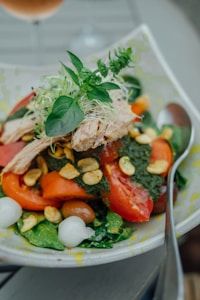 A colorful salad is presented on a plate, featuring fresh ingredients such as sliced tomatoes, leafy greens, shredded chicken, and sprouts. There are also small white onions, possibly pickled, and crunchy garnishes like cashews scattered on top. The salad is drizzled with a green pesto sauce, and a fork rests on the side of the plate.