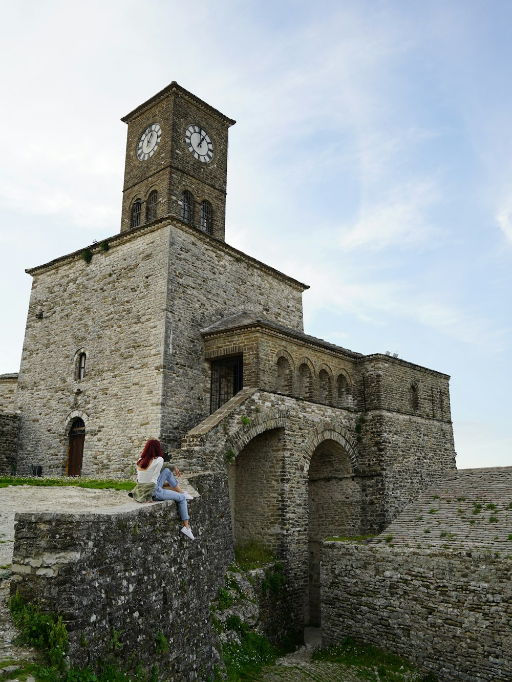 a woman sitting on a stone wall in front of a building with a clock tower