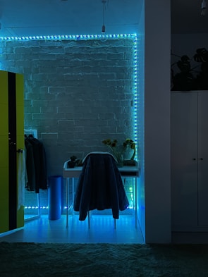 The image features a room with a modern aesthetic, highlighted by a brick wall illuminated with blue and green LED strip lights. A small desk is positioned against the wall with a chair draped in a coat. To the left, a bright yellow wardrobe contrasts against the dark, ambient lighting. A few plants and sunflowers in a vase are placed on the desk, adding a natural element to the decor.