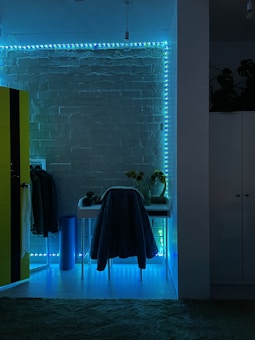 The image features a room with a modern aesthetic, highlighted by a brick wall illuminated with blue and green LED strip lights. A small desk is positioned against the wall with a chair draped in a coat. To the left, a bright yellow wardrobe contrasts against the dark, ambient lighting. A few plants and sunflowers in a vase are placed on the desk, adding a natural element to the decor.