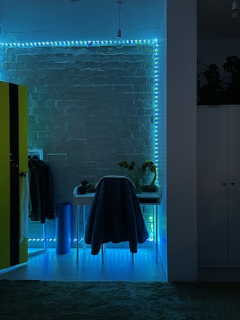 The image features a room with a modern aesthetic, highlighted by a brick wall illuminated with blue and green LED strip lights. A small desk is positioned against the wall with a chair draped in a coat. To the left, a bright yellow wardrobe contrasts against the dark, ambient lighting. A few plants and sunflowers in a vase are placed on the desk, adding a natural element to the decor.
